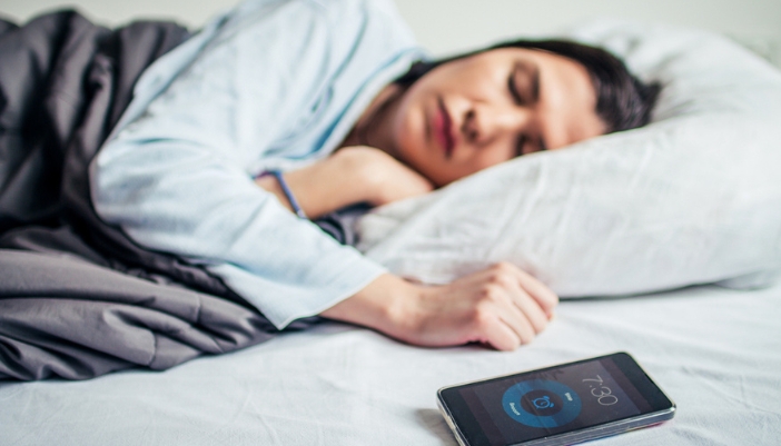 A woman sleeps as her phone is lit up to a sleep tracker app beside her on the mattress.