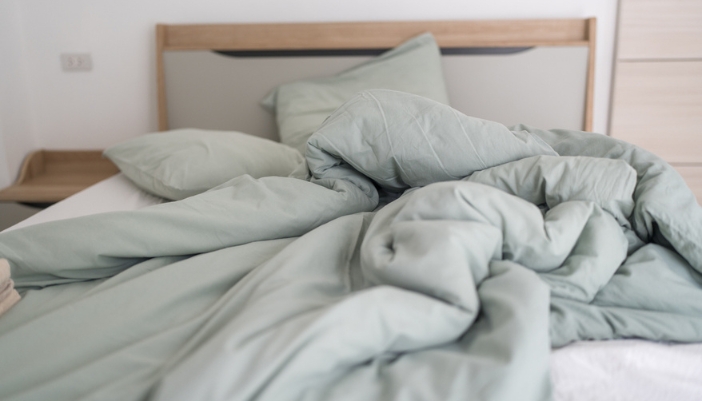 A pale green thick comforter sits mussed up on a bed at home.