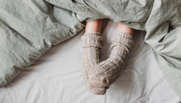 A pair of feet in cozy, thick tan sweater socks stick out from under covers on a bed.