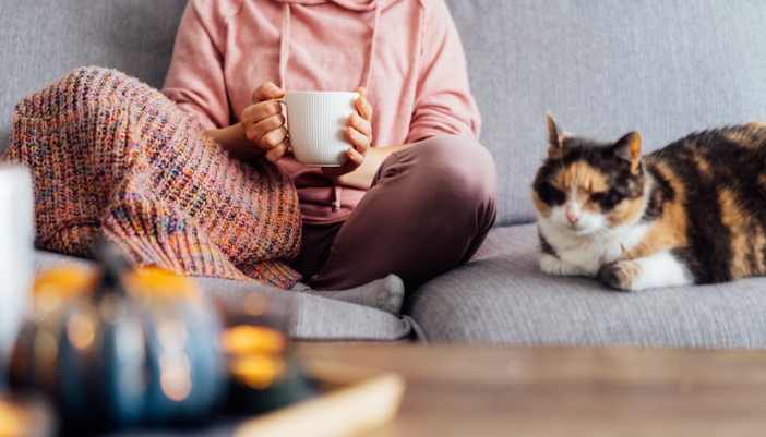 A woman sits on a gray couch next to her multi-colored fluffy cat and drinks tea.