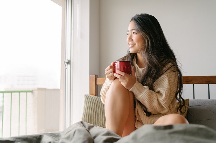 A young woman is drinking a cup of a hot drink in her bed and looking out of the widow in the morning.