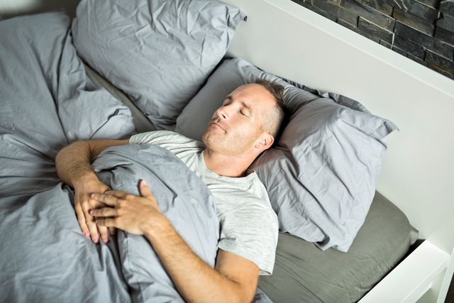 View of a man sleeping on his back on a bed with grey sheets