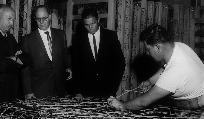 Black and white image of an older mattress factory with several men putting together a spring mattress. 
