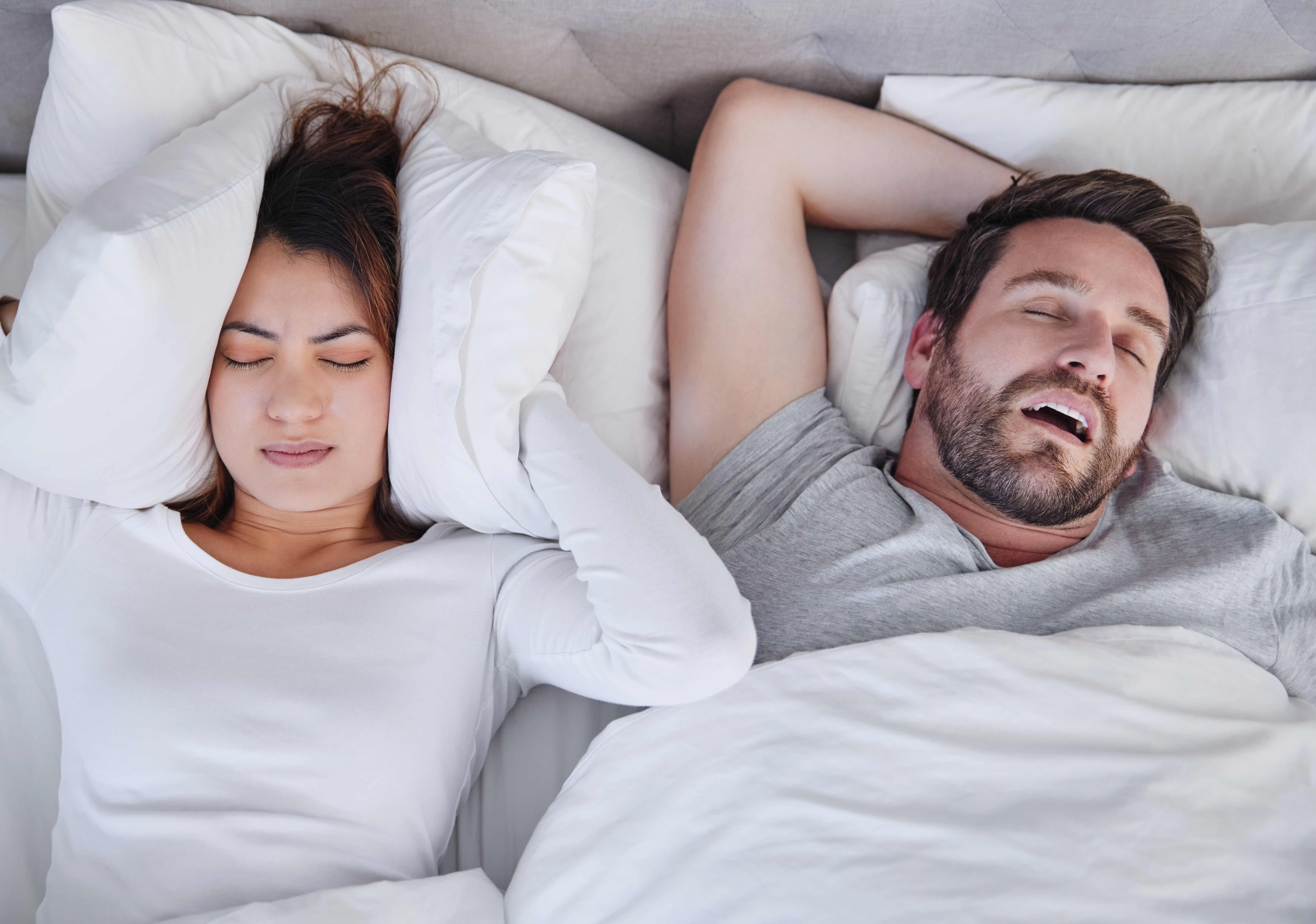 A woman covering her ears with a pillow as a man is snoring