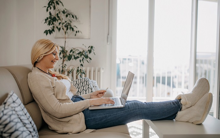 Middle aged woman sitting on a couch while online shopping on a laptop with her feet kicked up on a table. 