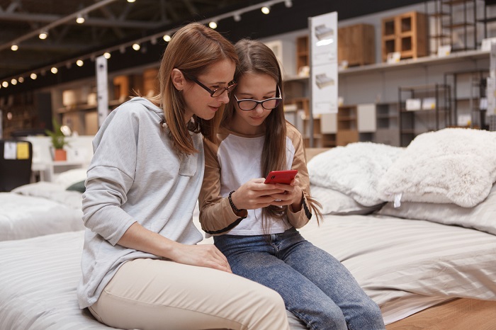 Mother and daughter sitting on a bed at furniture store, using smart phone together. 