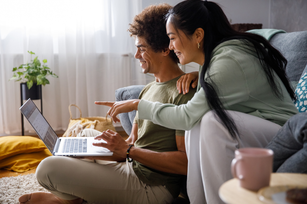 Shot of young man sitting on the floor and using laptop while his girlfriend is sitting on the sofa, behind him, and pointing at something on the screen.