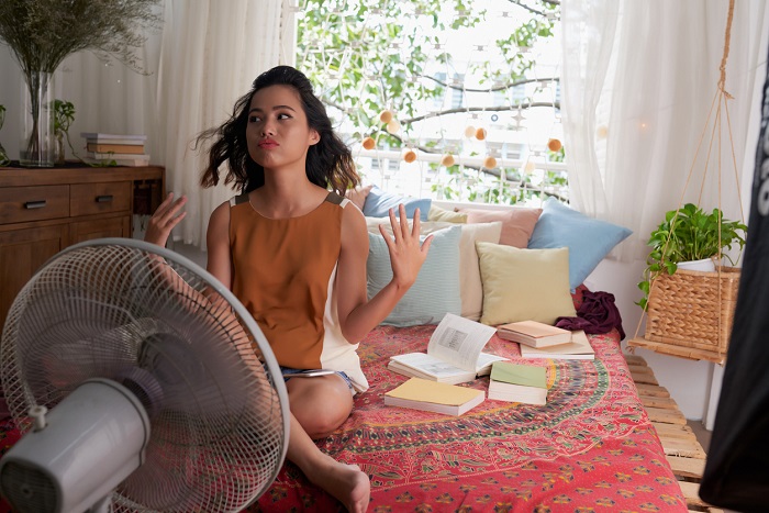 Girl cooling herself with big fan while sitting on bed.