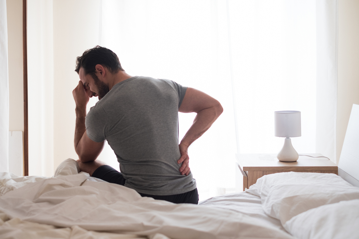 man sitting on side of bed holding his back in pain