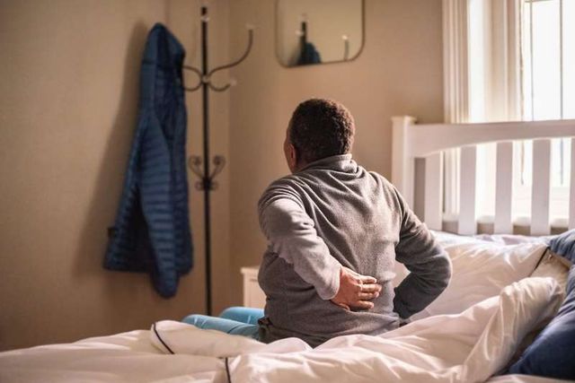 View of a woman waking up in a bedroom and holding her back while sitting on the side of her bed