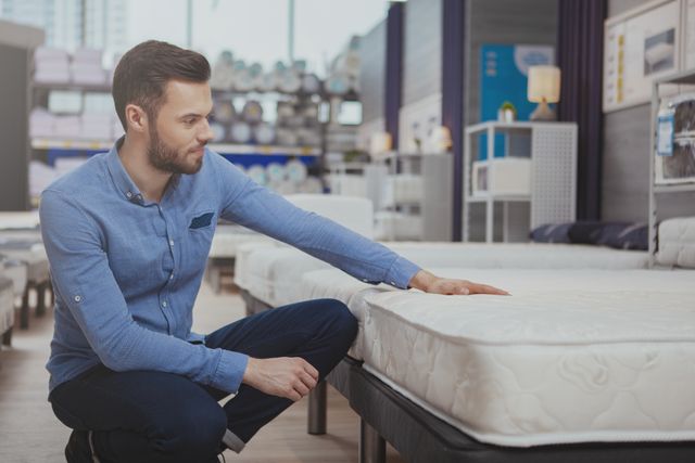 front view of a man kneeling and feeling a mattress in a mattress store