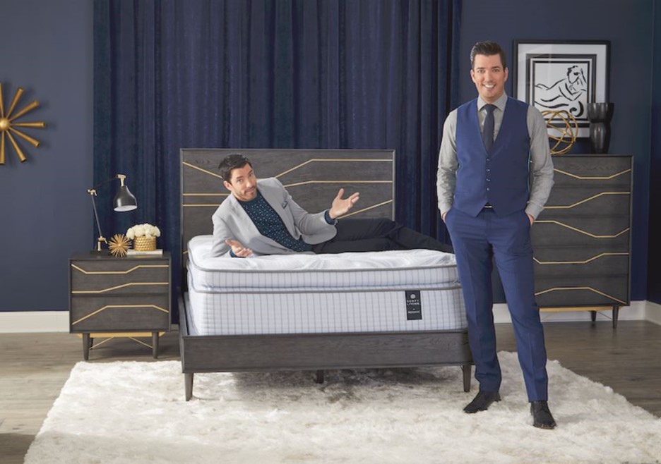 Drew and Jonathan Scott are shown in a dark blue bedroom showing off one of their mattress models