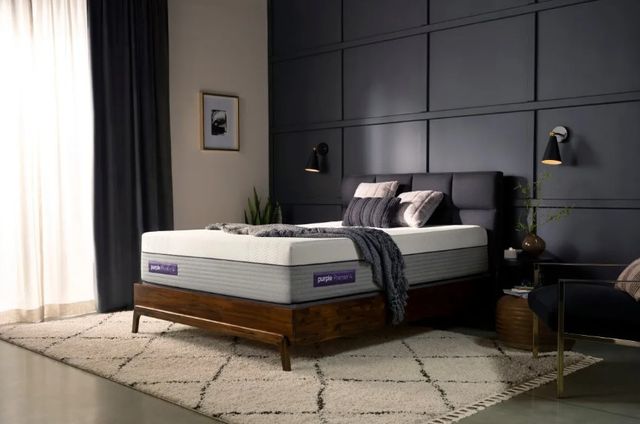 front view of a purple mattress in a contemporary bedroom with a grey throw and white and grey pilows