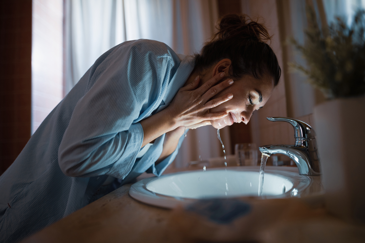young smiling woman washes her face over bathroom sink at night