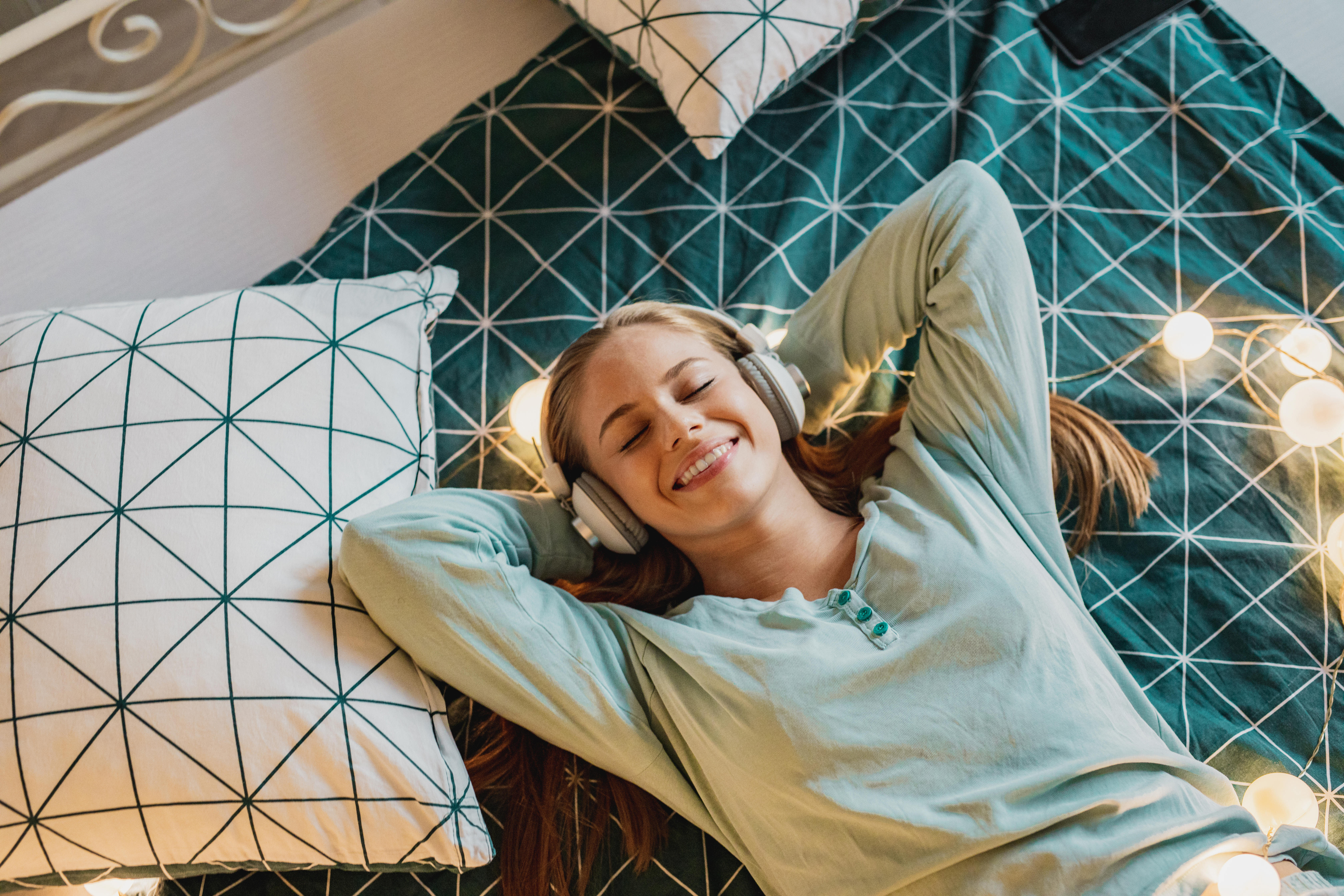 Young woman lying on teal and white bedding listens to music on headphones