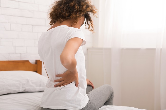 Woman sitting on the side of a bed holding lower back with one hand