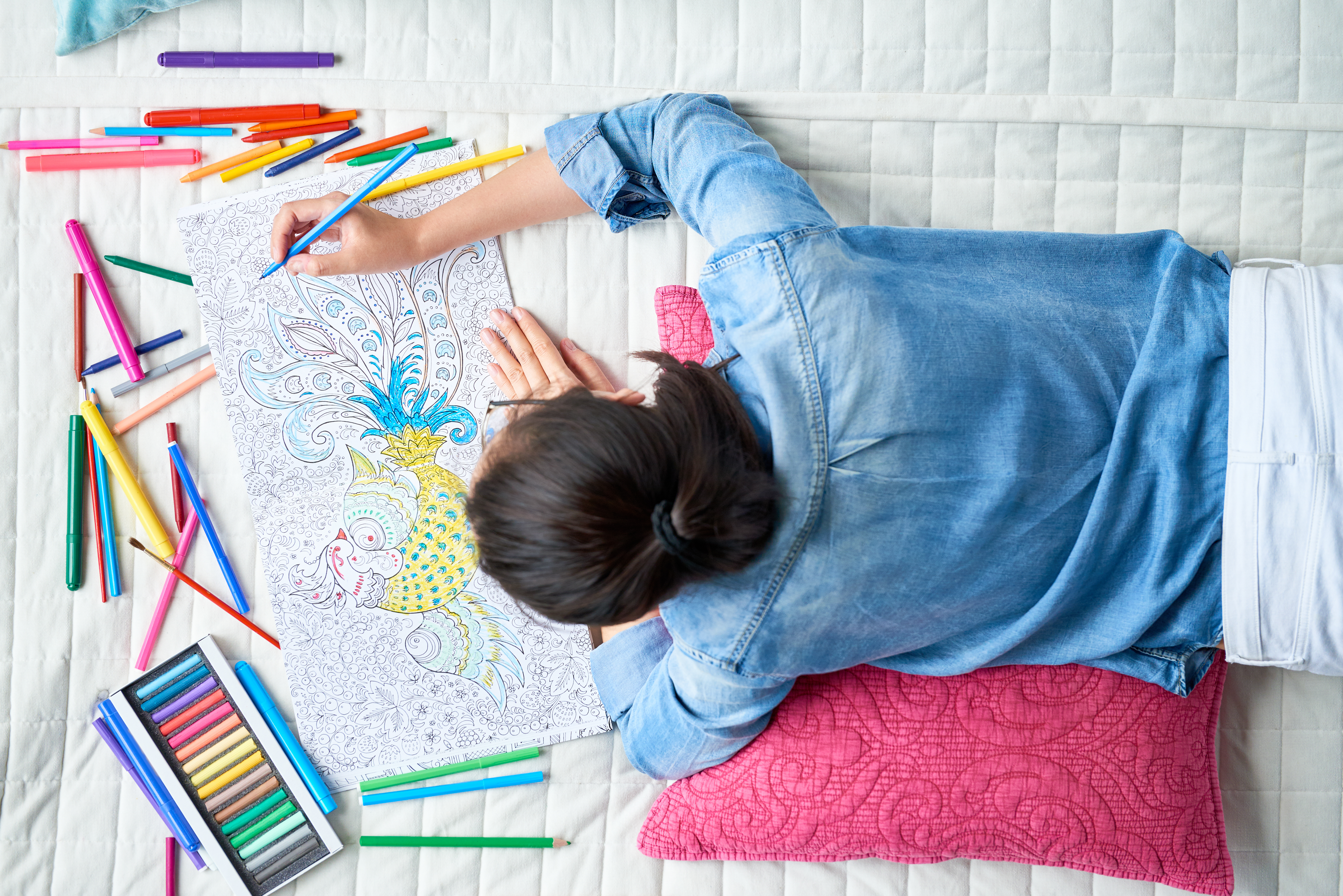 Young woman in jean jacket colors in an adult coloring book on a bed
