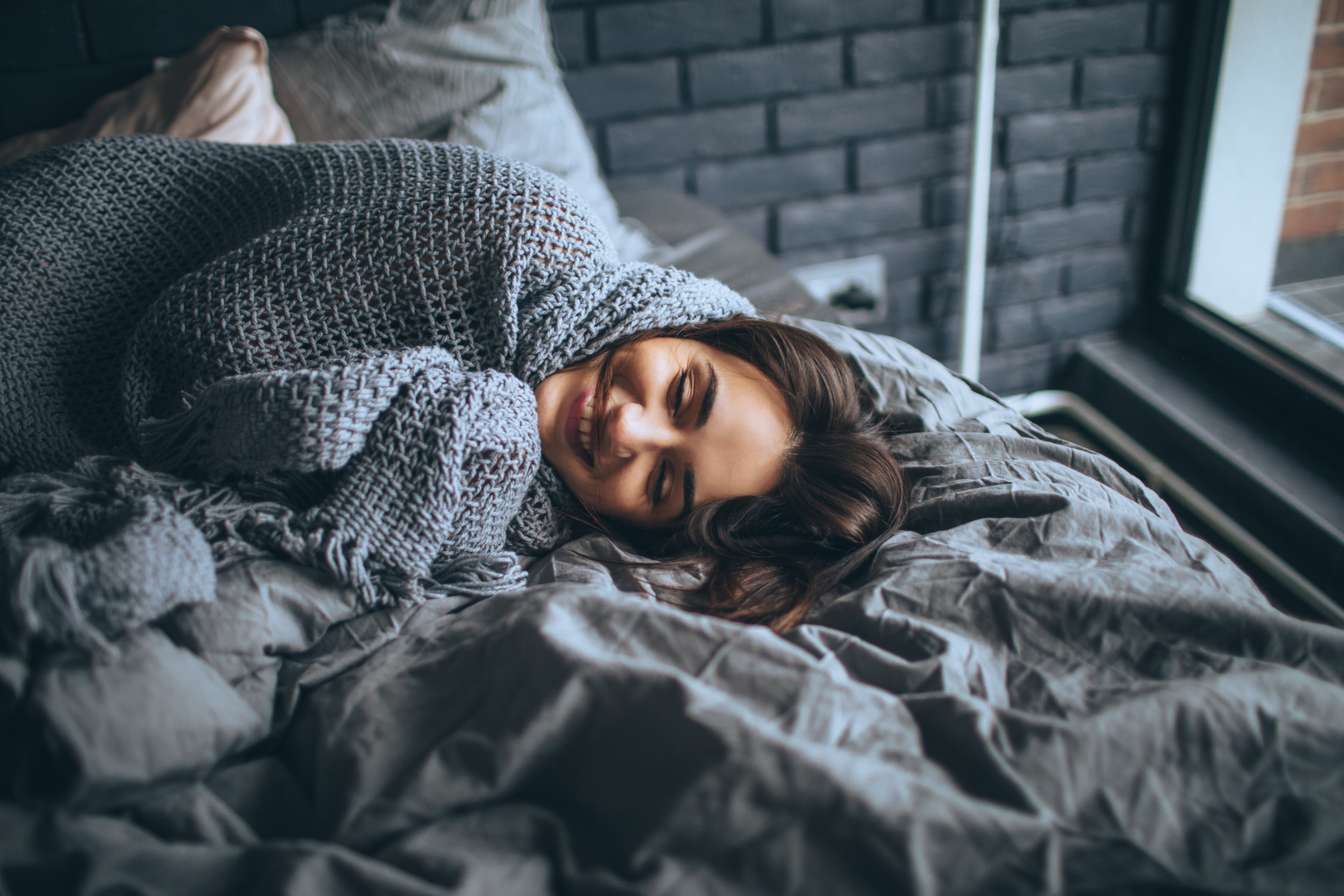young woman smiles as she wraps herself in gray knitted throw