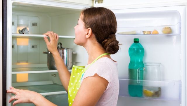Front view of a woman looking into the back of an empty refrigerator.