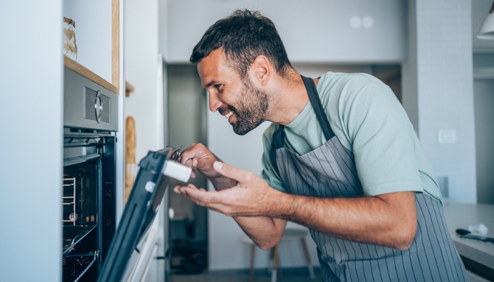 A man in an apron opens a stainless steel oven to look inside.
