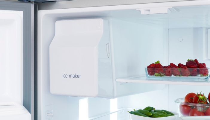 A fridge ice maker in a white fridge with bowls of fruit nearby.