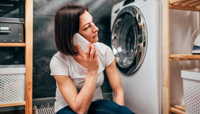 A woman sits in front of a white laundry appliance on a white phone.