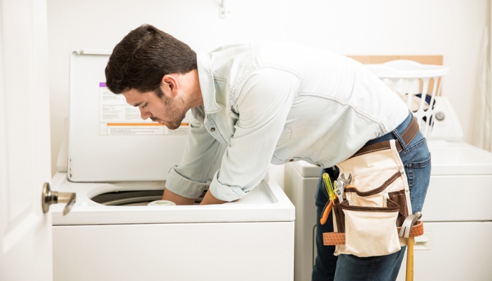 A young male handyman reaches into a laundry appliance to fix it.