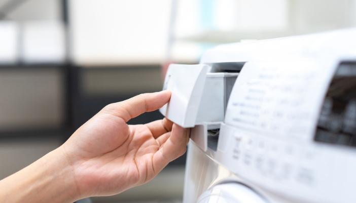 A hand opens a detergent dispenser on a laundry machine.