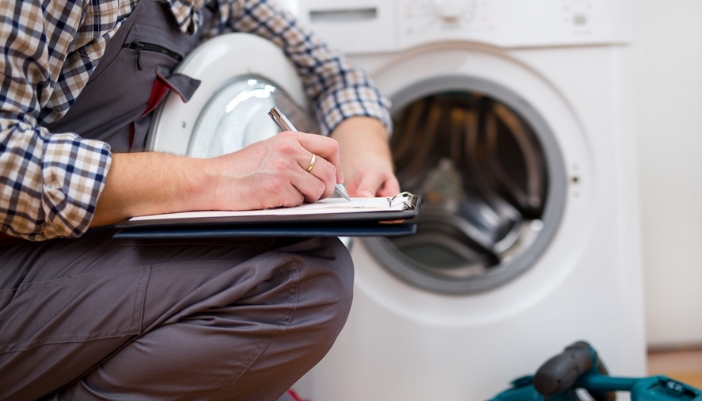 A repairman kneels in front of a washing machine filling out a sheet on a clipboard.