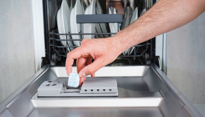 A male hand places a dishwasher detergent pod into an open dishwasher.