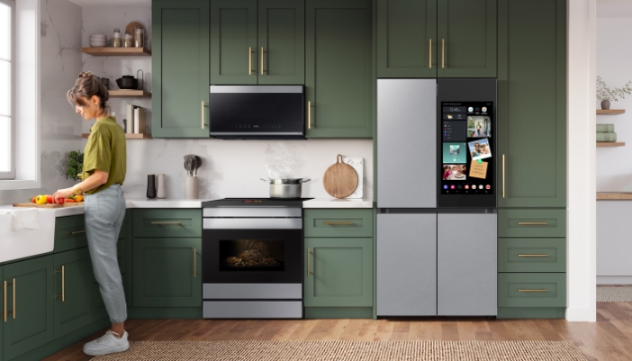 A woman works at a sink in her Samsung kitchen with green cabinets and gold accents.
