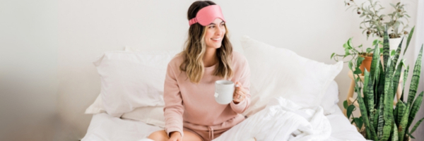 A woman sits on her bed with a sleep mask pushed up and a cup of coffee.