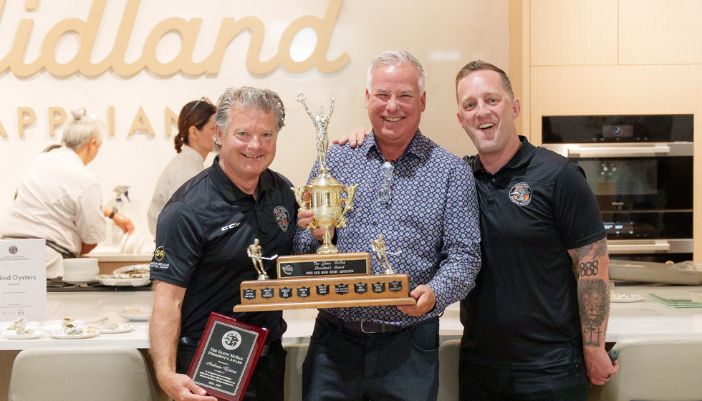 Three individuals stand in a Midland Appliance showroom kitchen, holding a trophy aloft, celebrating with smiles and excitement.