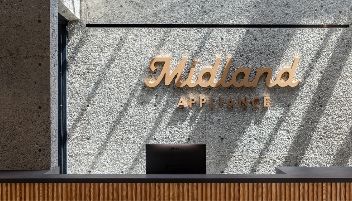 Modern reception area at Midland Appliance showroom featuring textured concrete walls, wood-accented desk, and gold signage under natural skylight.