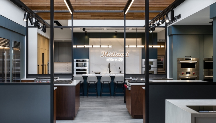 Modern kitchen showroom at Midland Appliance featuring luxury appliances, sleek cabinetry, and a central demonstration island under exposed wood ceiling.