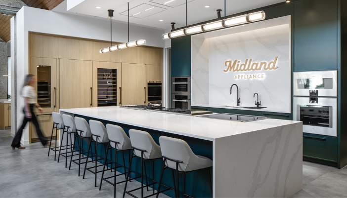 Modern kitchen showroom with a large marble island, bar stools, built-in ovens, and a sign reading "Midland Appliance" on the back wall. A person walks past on the left. 