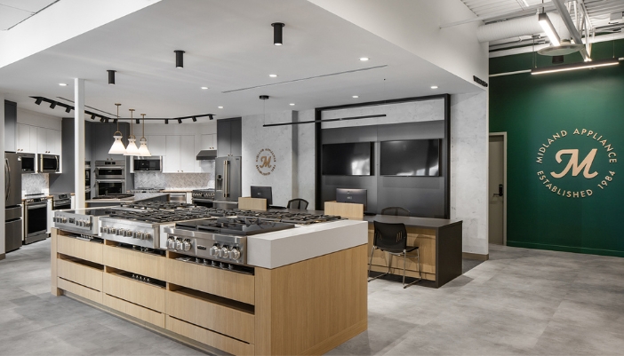 Modern appliance showroom featuring a central island with multiple cooktops, ovens along the walls, and a green wall with "Midland Appliance Established 1984" signage. 
