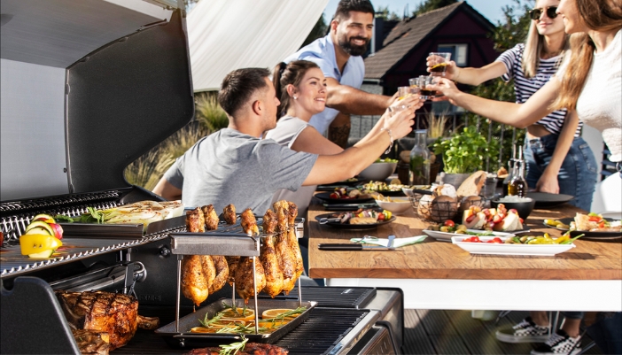 A group of friends sits next to a grill cooking and cheers glasses.