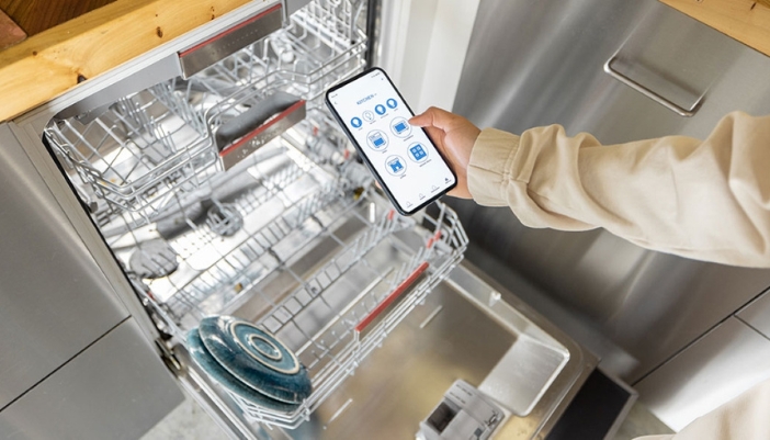 A hand uses a cell phone to control their smart dishwasher.
