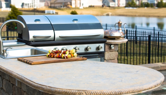 An outdoor kitchen featuring a sink and stainless steel grill with kebobs in front of it.