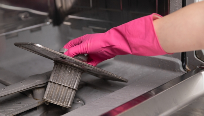 A hand in a pink rubber glove removes a filter from a dishwasher.