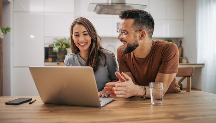 A couple sits at their kitchen table and uses a laptop together.