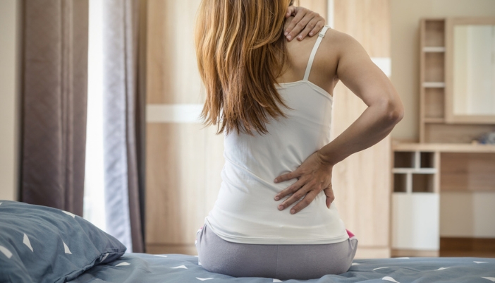 A woman sits on her bed rubbing her neck and back as if in pain.
