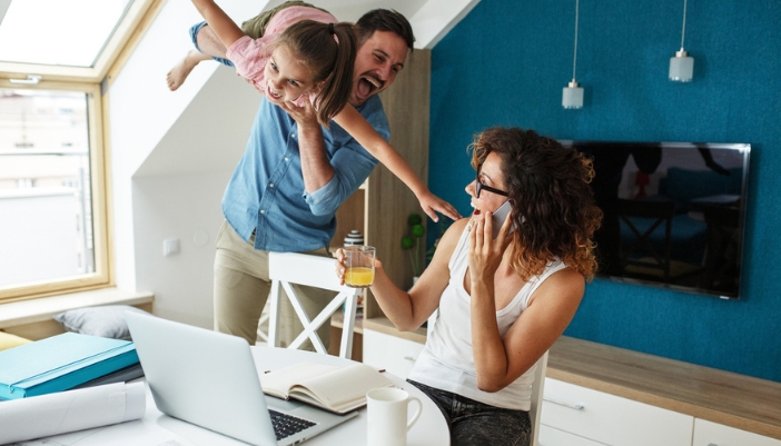 A mom works happily at a table at home while a father and daughter play nearby.