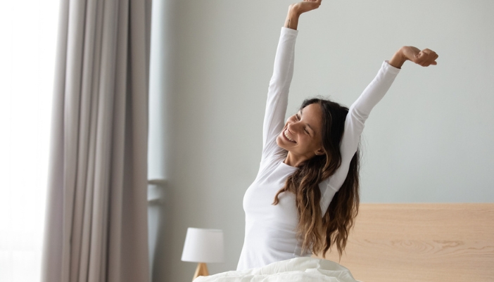 A woman wakes up happily and stretches her arms above her head.