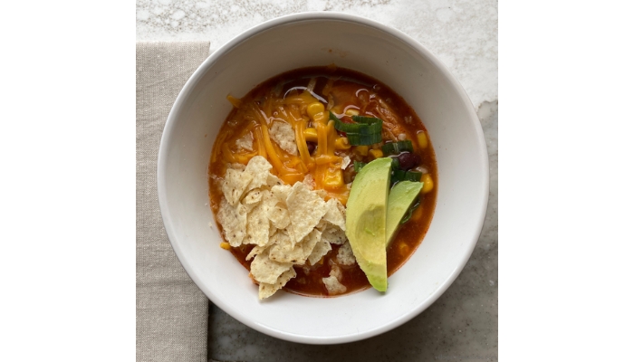 Overhead view image of chicken tortilla soup in white bowl topped with cheese, avocado slices, and tortilla chips