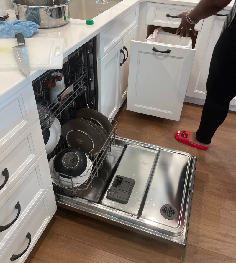 Dishwasher loaded with door open in white cabinet kitchen with woman opening waste basket cabinet pullout