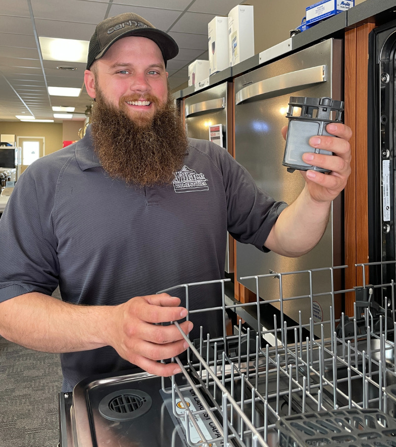 village service technician with beard and ball cap holding dishwasher filter near open display dishwasher