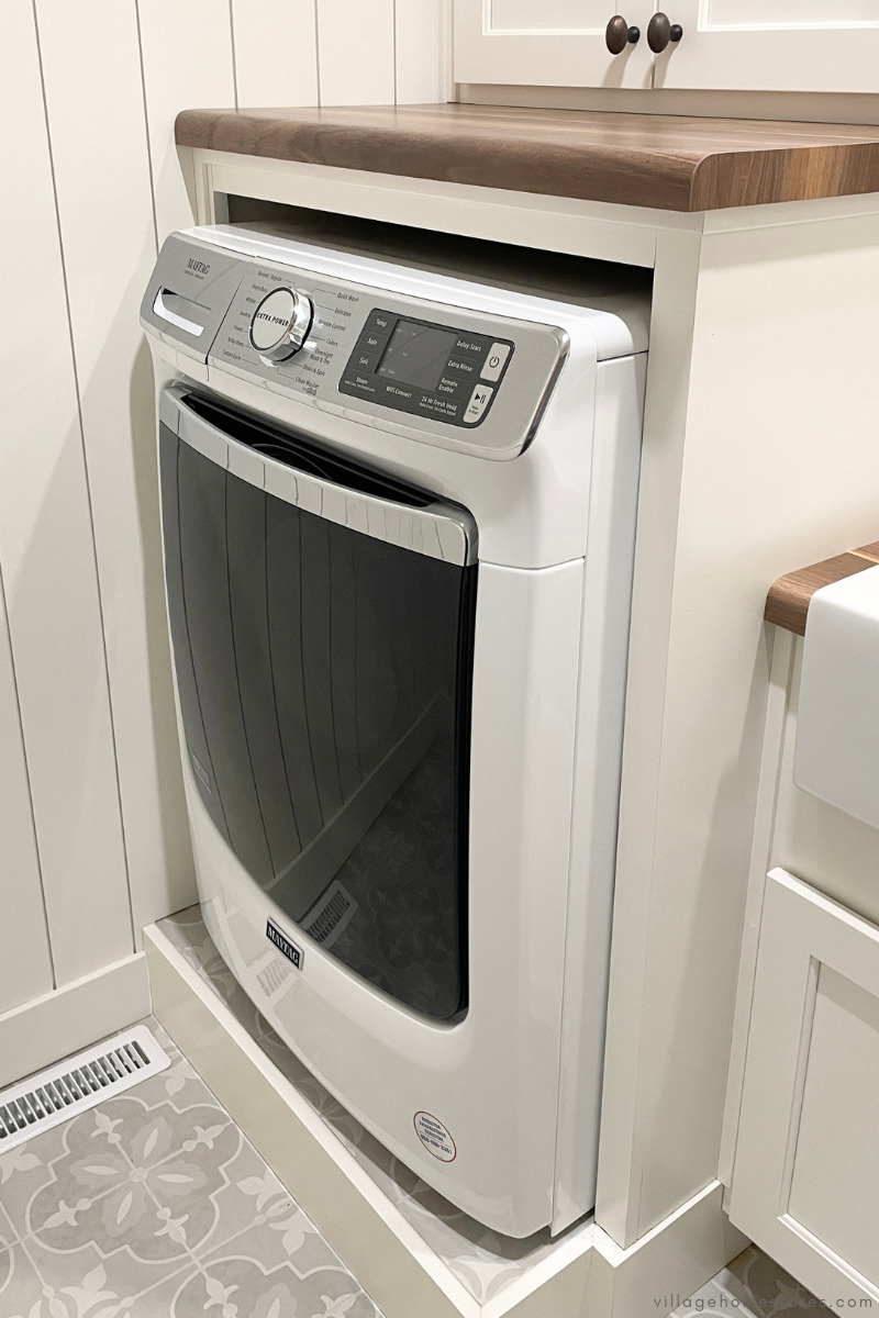 Alt. text: front load white maytag washing machine on tiled pedestal in laundry room with ivory painted cabinets and wood look countertops