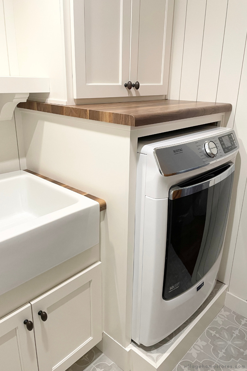 front load white maytag dryer on tiled pedestal in laundry room with ivory painted cabinets and wood look countertops.